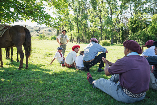 Group of smiling teenage Argentine gauchos relaxing in grass and talking with horses tied to nearby tree.