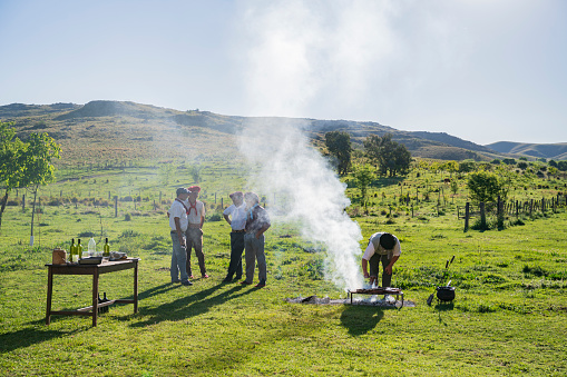 Young Argentine gauchos standing and talking in springtime sunshine while grill master checks meat being prepared for midday asado meal.