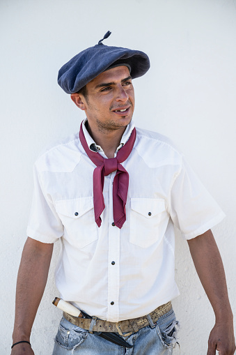 Close-up of young gaucho wearing traditional boina headwear, open collar shirt with scarf, facon tucked into waistband of jeans, and looking away from camera.