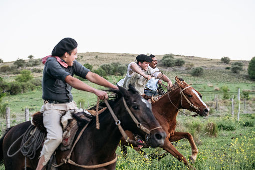 Side view of three Argentine gauchos wearing traditional clothing while galloping on horseback past camera in late afternoon.
