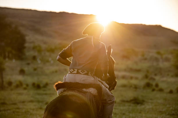 Young gaucho on horseback in late afternoon Rear view of teenage Argentine gaucho on horseback riding on grassland with sun setting over hills in background. gaucho stock pictures, royalty-free photos & images