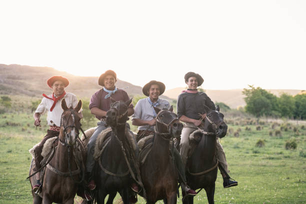 Four smiling teenage gauchos on horseback riding close together with sun setting over pampas hills in background.