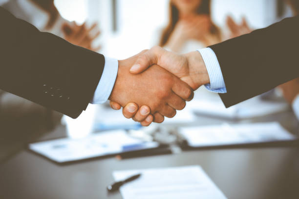 istock Business people shaking hands at meeting while theirs colleagues clapping and applauding. Group of unknown businessmen and women in modern white office. Success teamwork, partnership and handshake concept 1272540650