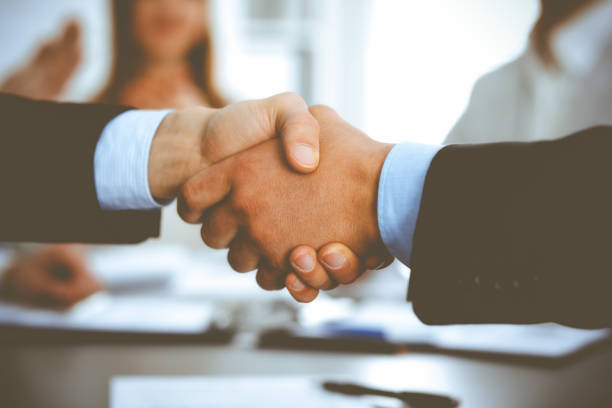 istock Business people shaking hands at meeting while theirs colleagues clapping and applauding. Group of unknown businessmen and women in modern white office. Success teamwork, partnership and handshake concept 1272540658