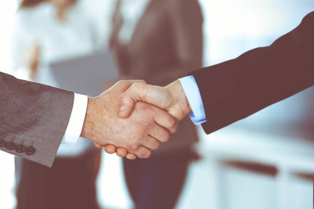 istock Business people shaking hands while standing with colleagues after meeting or negotiation, close-up. Group of unknown businessmen and women in modern office. Teamwork, partnership and handshake concept, toned picture 1272541707