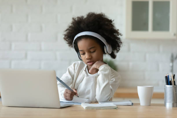 Mixed race child girl studying remotely online. Focused little 7s mixed race child girl wearing wireless headphones, listening educational online lecture on computer, studying remotely writing notes in copybook, e-learning distant education concept divergent movie review stock pictures, royalty-free photos & images