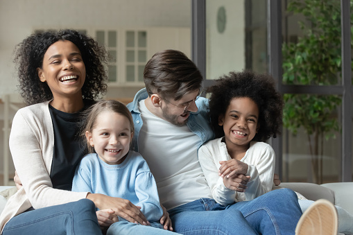 Happy emotional mixed race family relaxing together on comfortable couch indoors. Laughing multiracial couple enjoying watching television funny films with joyful small biracial children at home.