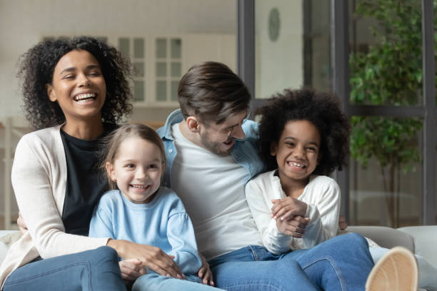 Laughing multiracial couple enjoying watching television funny films with kids. Happy emotional mixed race family relaxing together on comfortable couch indoors. Laughing multiracial couple enjoying watching television funny films with joyful small biracial children at home. different families stock pictures, royalty-free photos & images