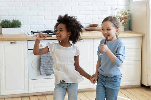 Funny adorable little mixed race preschool kids enjoying singing songs in kitchen utensils as microphones. Happy overjoyed small african ethnicity girl having fun with european stepsister or friend.