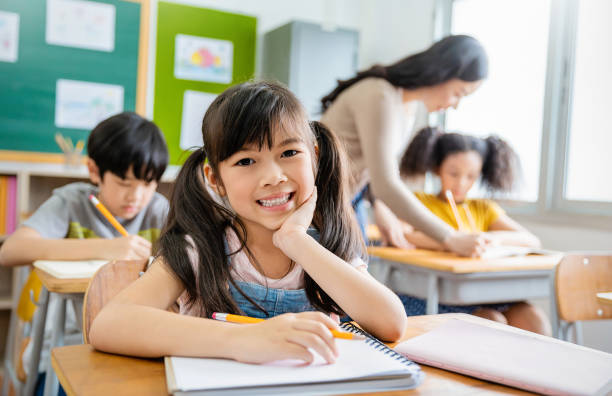 小学校の教室の机に書いている笑顔の小さな生徒の肖像画。小学校でテストをしている女子学生。教室でメモを書く子供たち。教育知識コンセプト - elementary student little boys classroom child ストックフォトと画像