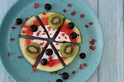 Stock photo showing elevated view of triangular slices of watermelon pizza fruit salad topped with yoghurt, kiwi, blueberries and pomegranate seeds, on a turquoise blue plate against a blue woodgrain background.