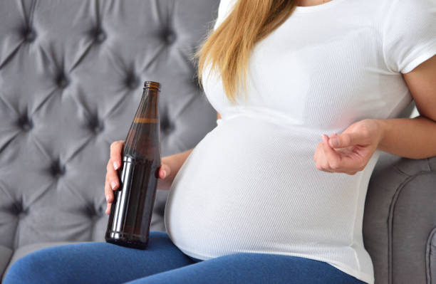 femme enceinte buvant la bière avec l’alcool tout en s’asseyant sur le divan - abdomen addiction adult alcohol photos et images de collection