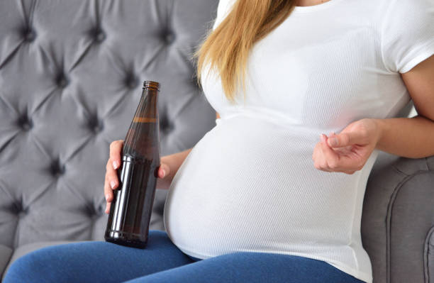 Pregnant woman drinking beer with alcohol while sitting on the couch stock photo