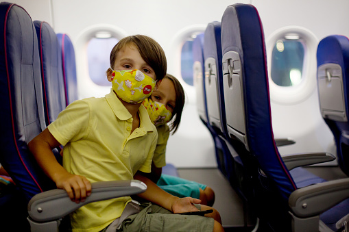 School boys, children boarding on the airplane, sitting and waiting for departure, wearing medical masks