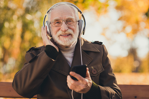 Photo of cheerful retired old grey haired grandpa feel young street sit bench listen youth song music big earphones follow grandchildren, trends positive emotions wear glasses jacket outside