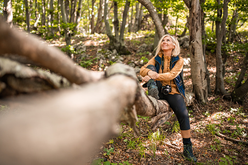 Adult caucasian cheerful woman with a camera around her neck sitting on a tree trunk in woods and looking up with a smile on her face.