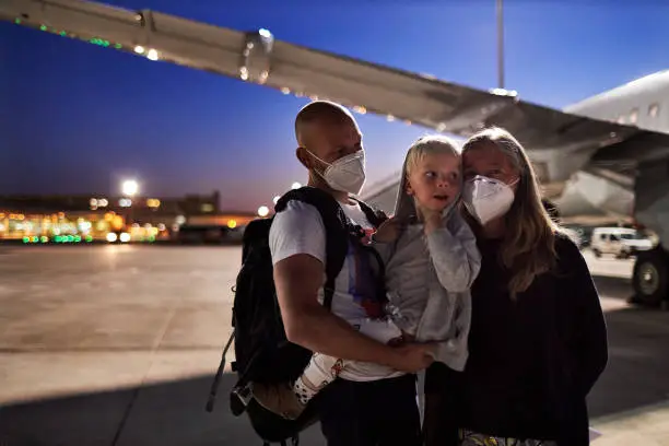 A senior woman is standing with a young father who is wearing a surgical mask or face mask and has his son in his arms in the dark on the tarmac in front of the plane before they go on vacation, high-resolution photographed with copy space