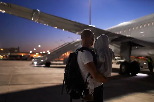 A young father wearing a surgical mask or face mask has his son in his arms and is standing in the dark on the tarmac in front of the plane before they go on vacation, high-resolution photographed with copy space