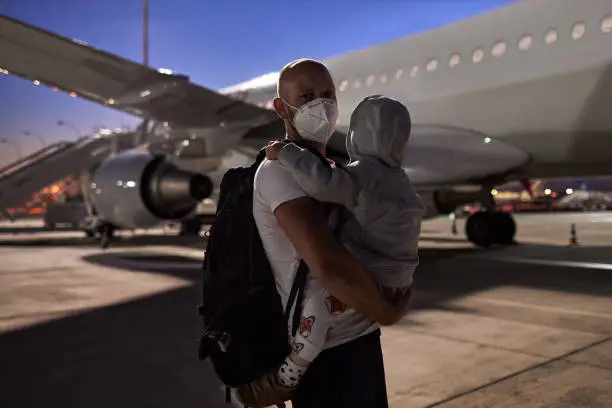 A young father wearing a surgical mask or face mask has his son in his arms and is standing in the dark on the tarmac in front of the plane before they go on vacation, high-resolution photographed with copy space