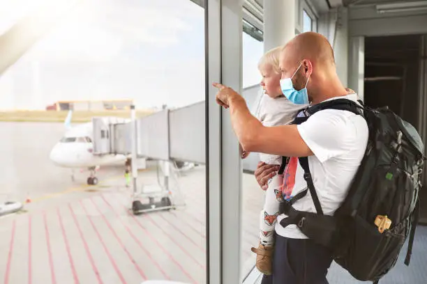 A young father with a surgical mask or face mask has his son in his arms and shows him the planes through a window at the airport before they go on vacation, photographed in high resolution with copy space