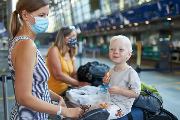 A woman with blond hair and a surgical mask or face mask is standing with a suitcase cart full of luggage on which her little son is sitting and eating an apple at the airport before they go on vacation, high-resolution photographed with copy space