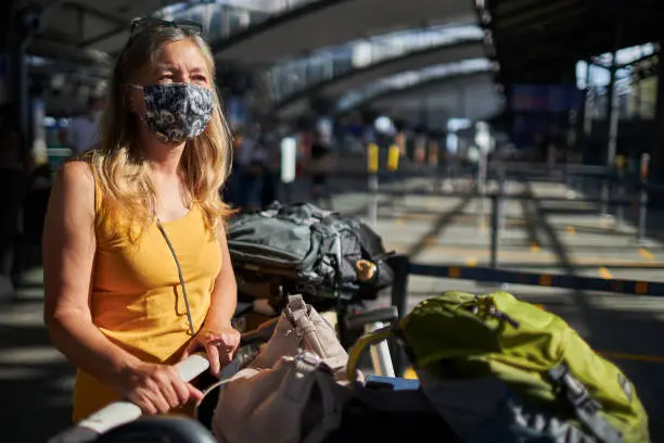 A senior woman with long hair and a surgical mask or face mask stands thoughtfully with a luggage cart full of luggage at the airport before she goes on vacation, high-resolution photographed with copy space