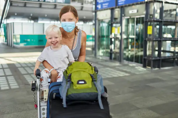 A young mother pushes a suitcase trolley on which her little son is sitting through the airport and wears a surgical mask or face mask, photographed in high resolution with copy space