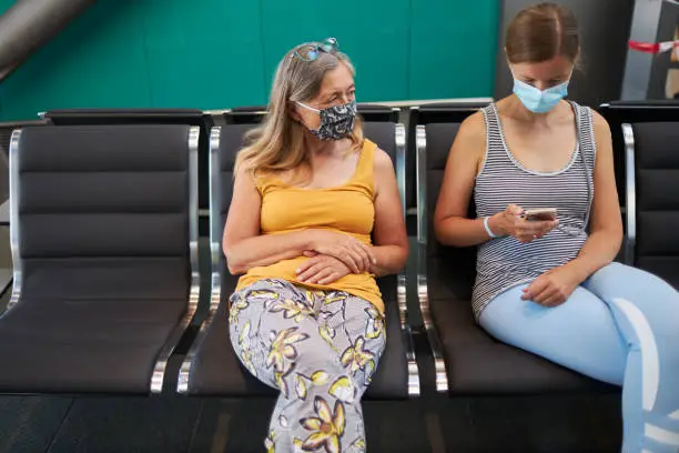 Two women with face masks or surgical masks sit next to each other at the airport while the younger one looks into a smartphone or cell phone, photographed in high resolution with copy space