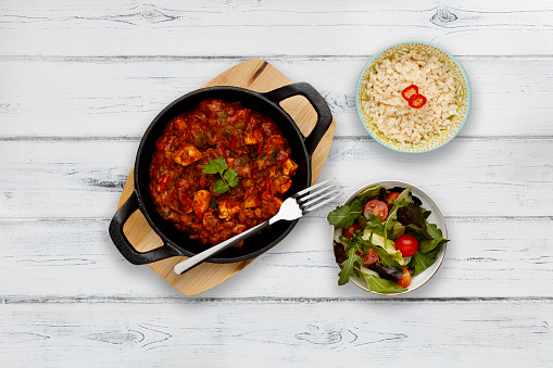A traditional blati dish filled with delicious balti chicken curry, garnished with a sprig of corriander, on a wooden serving board, with a side dish of rice and a bowl of salad, on a rustic white wooden background.