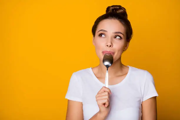 Portrait of positive inspired girl hipster lick lips spoon look copyspace, feel interested about what she will eat breakfast wear white t-shirt isolated over bright shine yellow color background