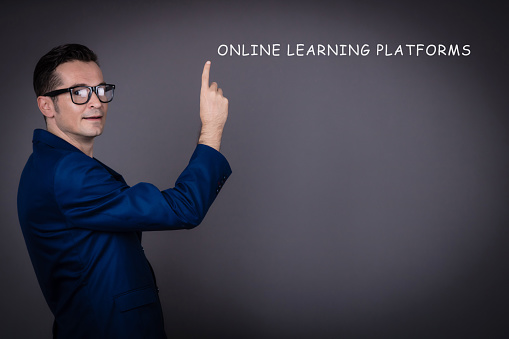Mid adult teacher holding a class in front of gray background and pointing at online platforms. Copy space.
