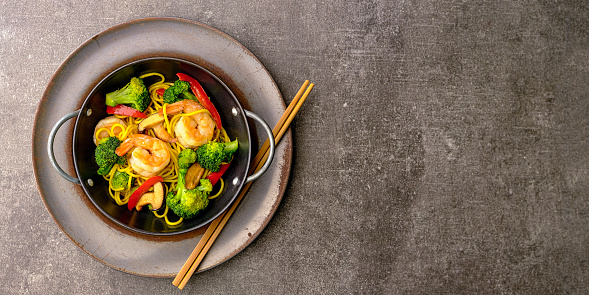 Delicious colorful stir-fried prawn and vegetable noodles in a personal serving dish wok with chopsticks resting on the side of the wok on a grungy grey background.