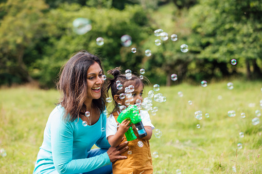 A medium shot of a Pakistani, mid adult mum and her young, mixed race son playing with a bubble gun in a woodland forest field in Northumberland, Northeastern England during the Covid-19 pandemic. The son is holding the bubble gun while his mother is helping him to use it.