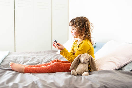 Side view little girl sitting on bed watching movie on phone