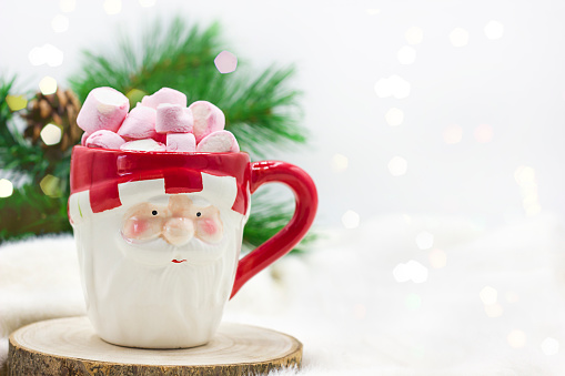 a cup in the form of santa claus with a hot drink and marshmallows standing on a wooden stand on a white background next to a green christmas tree branch. Space for text. Cozy winter concept