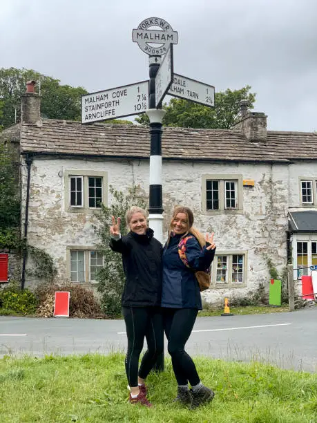 Two friends standing at either side of traditional cast iron road direction sign doing a peace sign in the village of Malham, Yorkshire.