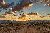 A dirt road leading to distant mountains where the sun has just set behind with colorful clouds