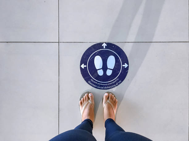 Directly above shot of an Asian young woman feet standing behind a safety floor marking sticker to keep social distance while queueing at the airport during Covid 19 pandemic