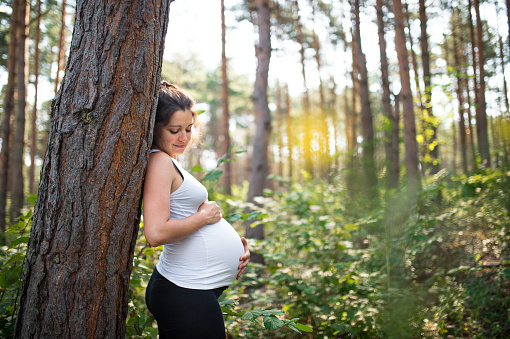 Side view portrait of happy pregnant woman outdoors in nature, touching her belly.