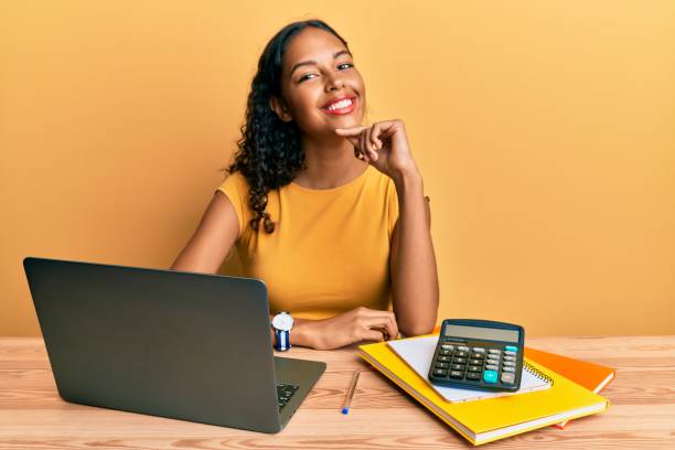 młoda afroamerykanka pracująca w biurze z laptopem i kalkulatorem patrząca pewnie na kamerę uśmiechnięta ze skrzyżowanymi ramionami i ręką podniesioną na brodzie. pozytywne myślenie. - arms crossed zdjęcia i obrazy z banku zdjęć