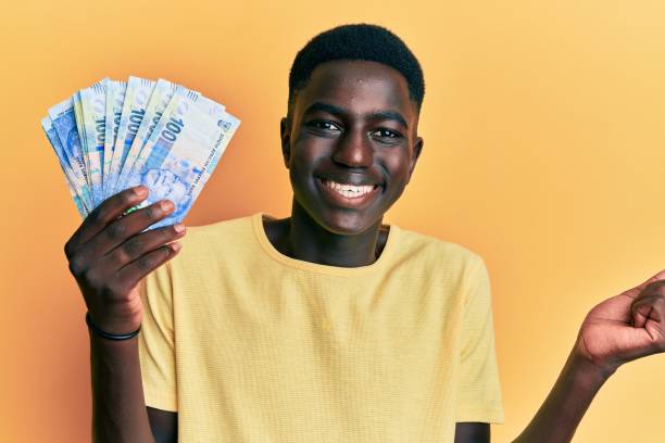 Young african american man holding south african rand banknotes screaming proud, celebrating victory and success very excited with raised arm Young african american man holding south african rand banknotes screaming proud, celebrating victory and success very excited with raised arm african currency stock pictures, royalty-free photos & images