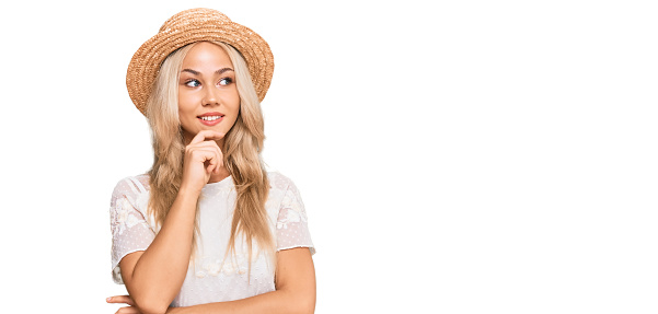 Young blonde girl wearing summer hat with hand on chin thinking about question, pensive expression. smiling with thoughtful face. doubt concept.