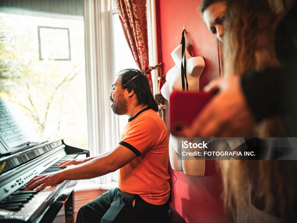 Man playing piano and singing Man playing piano and singing while his female band member is recording him on the phone beside him. Interior of home by the window. Recording Studio Stock Photo