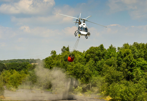 Pontypridd, Wales - July 2018: Helicopter in flight carrying a large water carrier bucket slung underneath to fight a large grass fire in South Wales. The rotor blades have churned up a dust cloud.