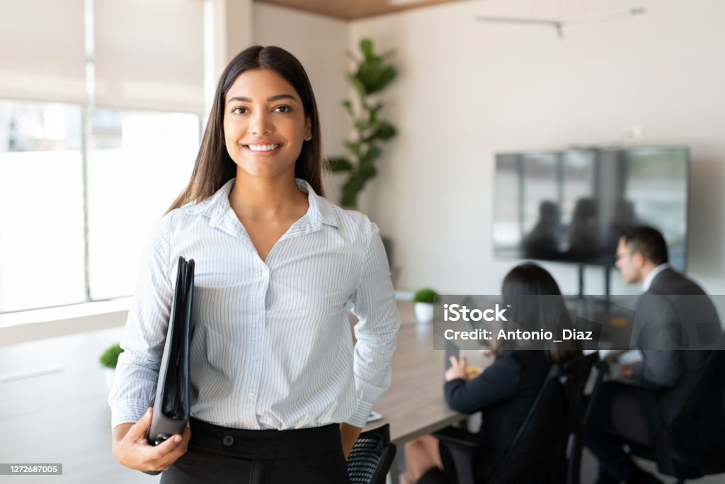 Hispanic female business professional in office boardroom Portrait of hispanic businesswoman with a file standing in meeting room with colleagues disucssing in background Office Stock Photo