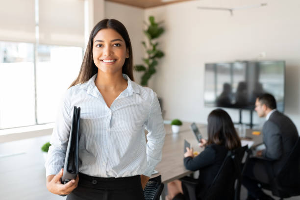 Hispanic female business professional in office boardroom Portrait of hispanic businesswoman with a file standing in meeting room with colleagues disucssing in background colleague photos stock pictures, royalty-free photos & images