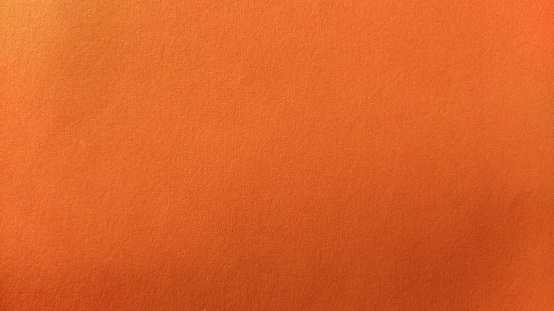 A sheet of thick paper of bright orange color. Close-up. Background intense shade. Natural lateral lighting. Fine cardboard or paper texture. Light gradient in degree of illumination.