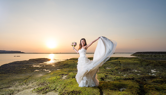 A young Japanese woman in a wedding dress on the beach at sunset in Okinawa, Japan. She is lifting up her wedding dress.