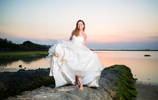 A young Japanese woman in a wedding dress on the beach at sunset in Okinawa, Japan. She is lifting up her wedding dress as she walks on the rocks.