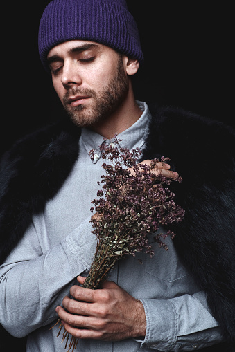Portrait of young attractive male with beard in fur coat and purple hat holding bouquet of flowers with his eyes closed